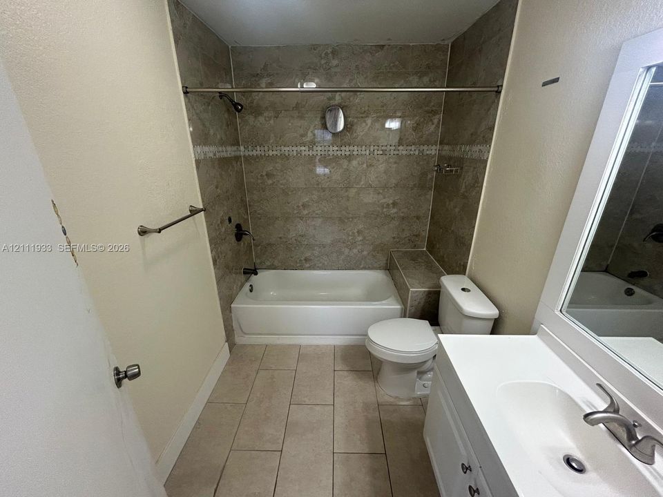Bathroom featuring stone-finish tile shower surround with integrated bench, white tub, neutral floor tile, and vanity with built-in mirror and integrated sink