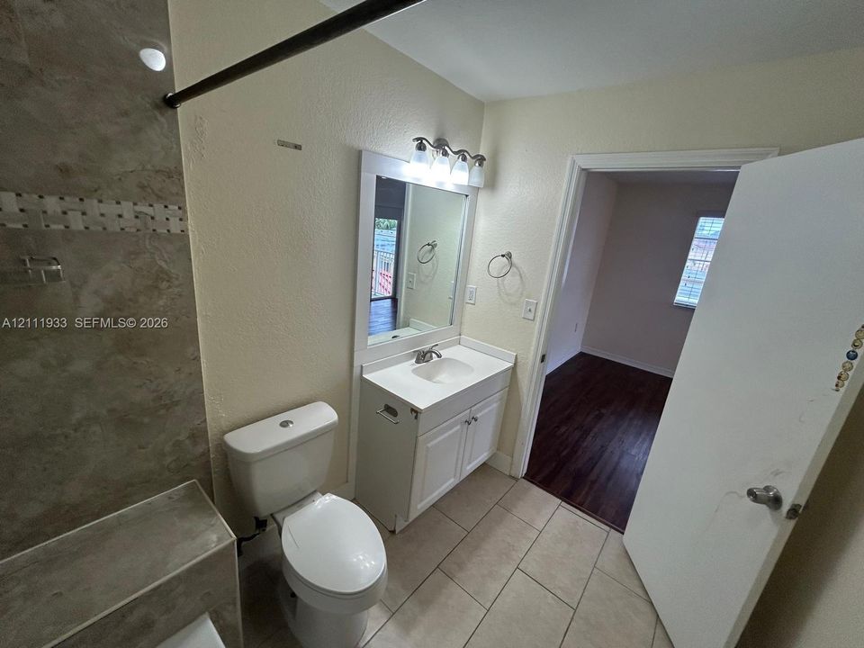 Bathroom featuring tile flooring, white cabinetry with a solid surface countertop, rectangular undermount sink, framed vanity mirror with integrated lighting, and tile shower surround