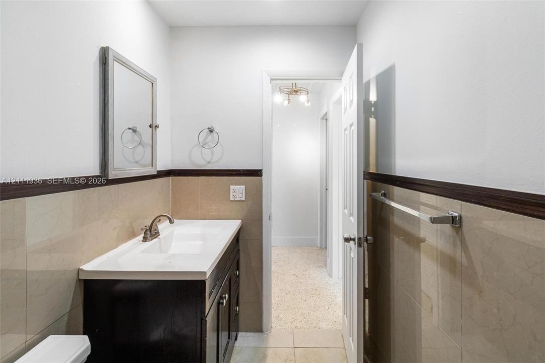 Bathroom featuring a vanity with an integrated basin, wall-mounted mirror, polished metal hardware, and tiled wall wainscoting with a dark wood-finish rail