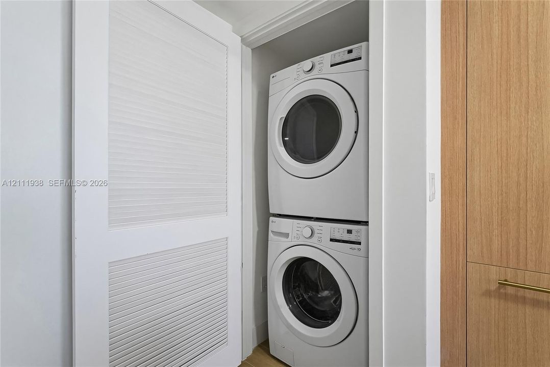 Stacked laundry unit featuring white appliances, enclosed by white louvered bifold doors and wood-finish cabinetry