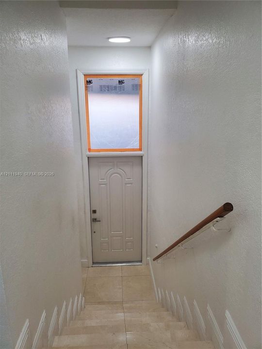 Staircase with tile-covered steps, white textured walls, and a wood-finish handrail with white metal brackets, featuring a single-panel door with a transom window and a flush-mount ceiling light