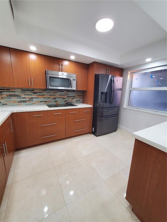 Kitchen featuring wood-finish cabinetry, solid surface countertops, multi-toned glass tile backsplash, large-format tile flooring, and stainless steel appliances