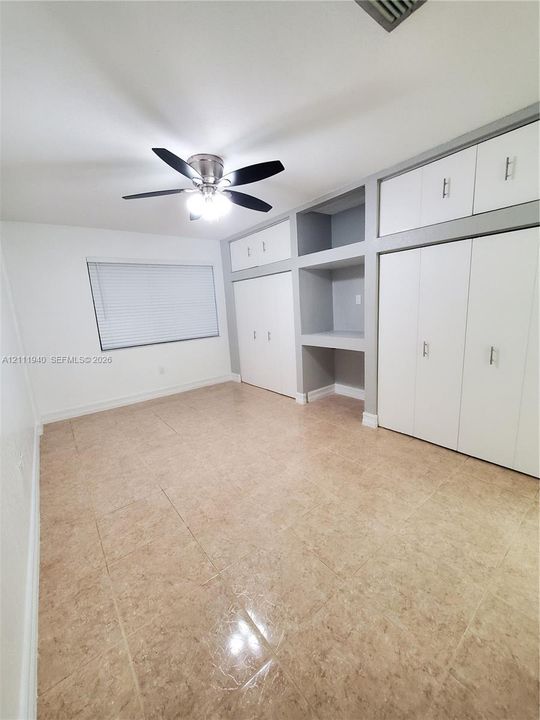 Room interior featuring tile flooring, ceiling fan with integrated lighting, window with horizontal blinds, and extensive built-in floor-to-ceiling storage cabinetry with shelving