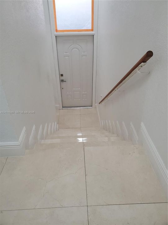Staircase featuring large-format tile flooring and a wall-mounted wooden handrail, terminating at an arched-panel exterior door with an overhead transom window