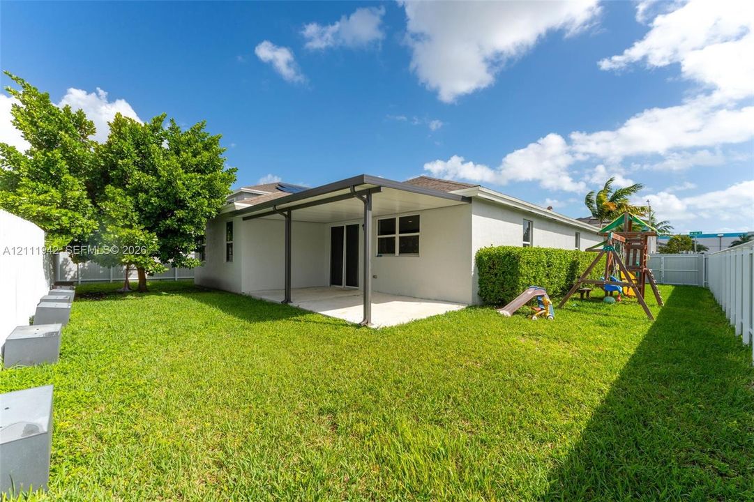 Rear exterior space featuring a covered patio, manicured lawn, mature trees, and perimeter white vinyl fencing