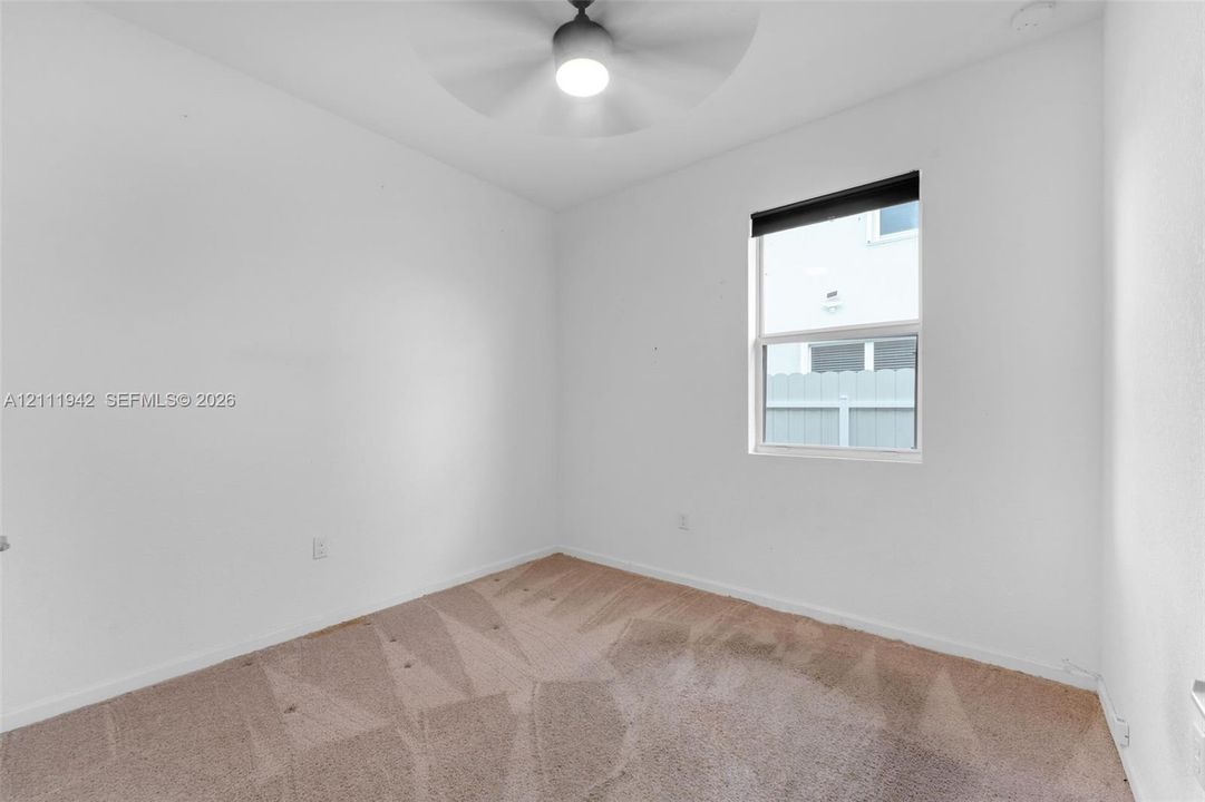 Neutral-toned carpeting, matte black ceiling fan with integrated lighting, and single-pane window with dark roller shade