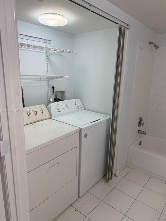 Laundry closet featuring wire shelving and overhead lighting, with white tile flooring and a bathtub enclosure with a wall-mounted showerhead