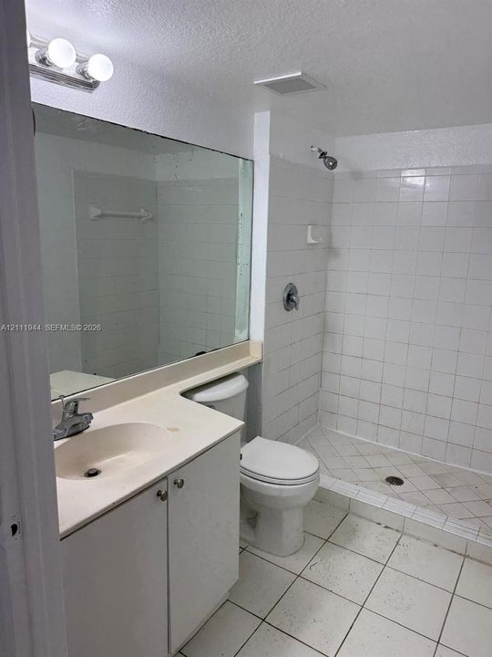 Bathroom featuring a vanity with integrated sink, large wall-mounted mirror, three-bulb vanity light, toilet, and a walk-in shower with tiled walls and flooring