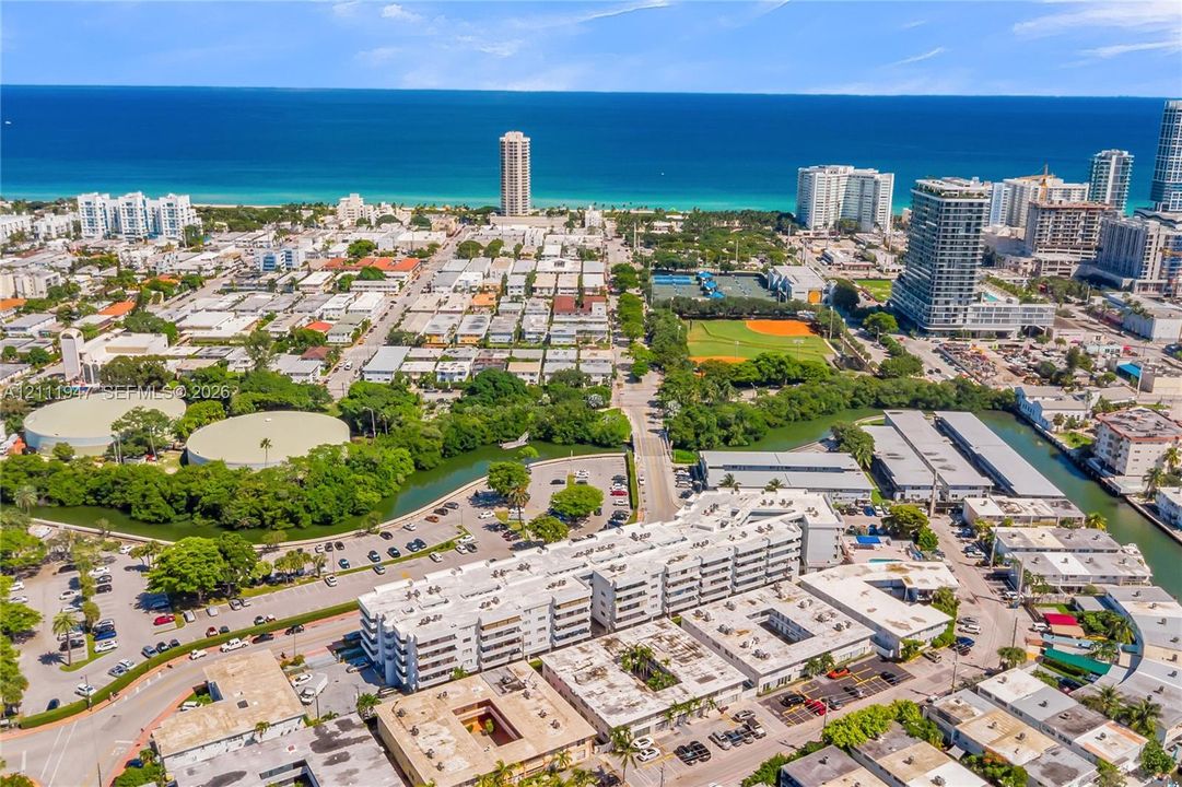 Aerial perspective of urban landscape featuring multi-story residential structures, proximity to coastal shoreline, paved surface parking, and manicured green spaces
