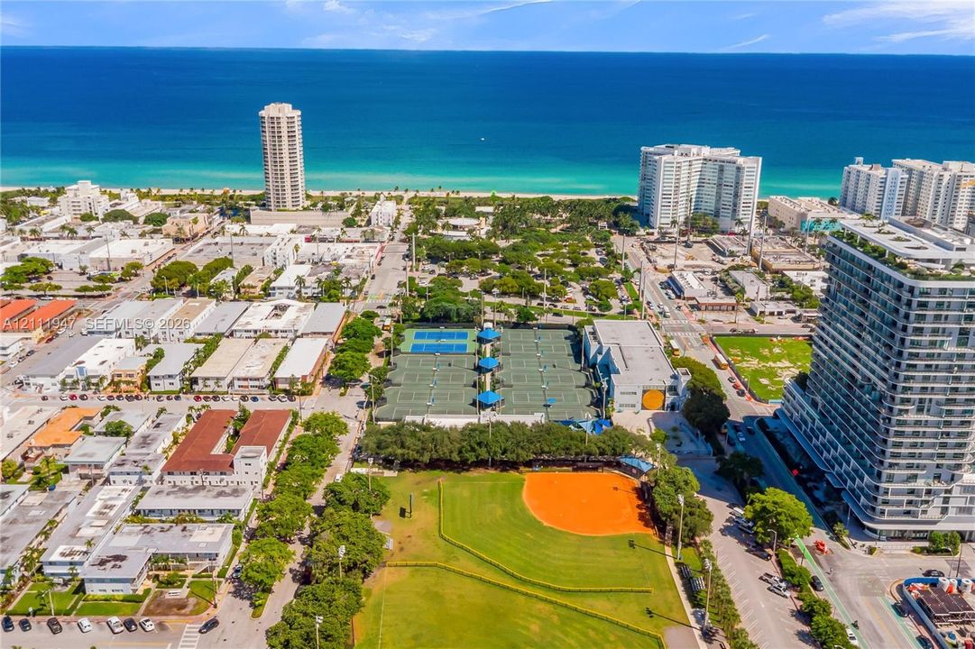 Aerial coastal setting featuring a public park with multiple tennis courts, a baseball field, mature greenery, and proximity to the oceanfront