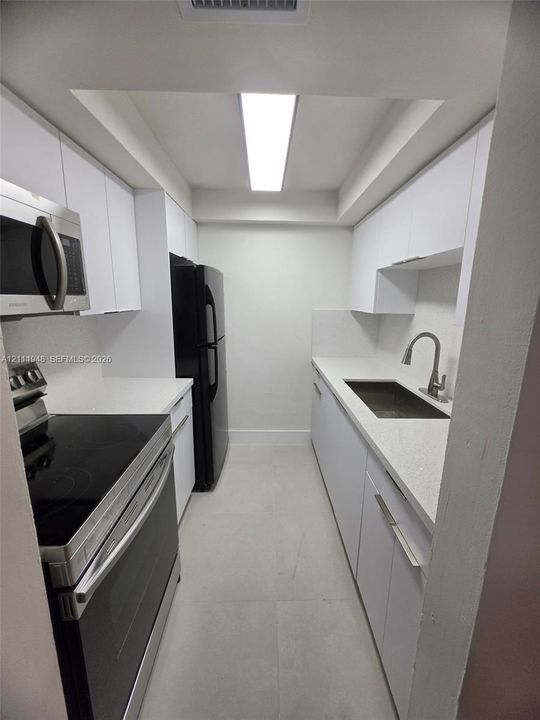 Galley kitchen featuring white flat-panel cabinetry, light-toned stone-surface countertops, large-format tile flooring, a deep undermount sink with a gooseneck faucet, and stainless-steel appliances