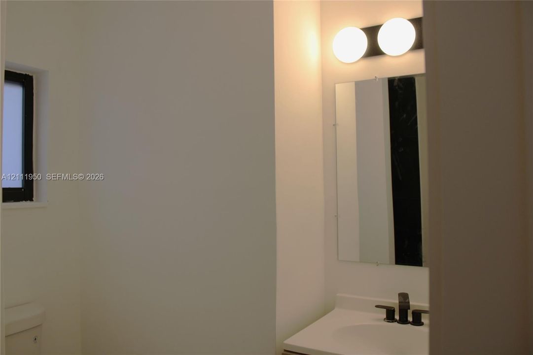 Wall-mounted globe vanity lighting, rectangular frameless mirror, white composite vanity top with an integrated sink, and a dark finish centerset faucet