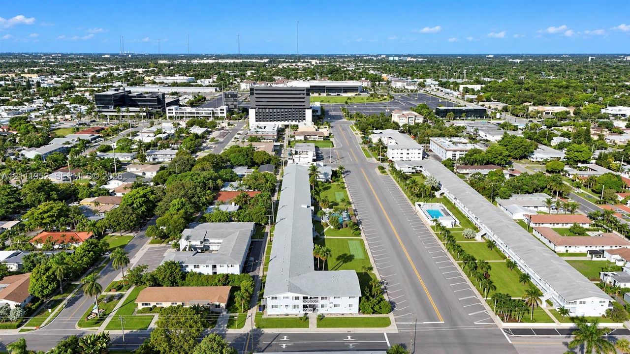Aerial perspective of a multi-story residential building featuring a light-colored exterior finish, a flat roofline, manicured perimeter landscaping, and dedicated surface parking