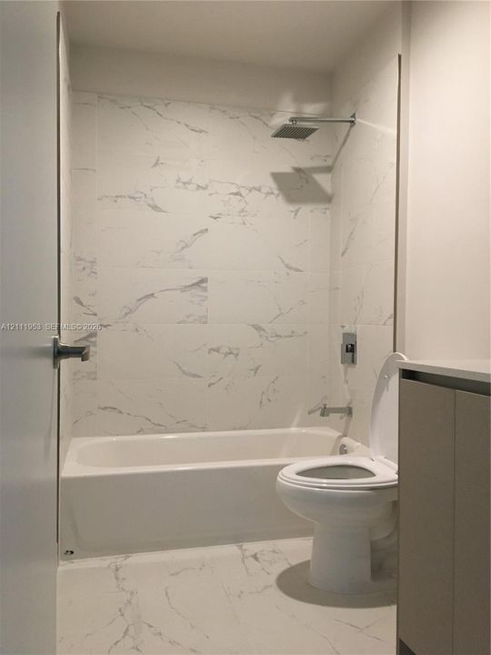 Bathroom featuring full-height large-format stone-patterned tile surround, built-in bathtub, rainfall showerhead, wall-mounted faucet controls, and solid-surface vanity cabinetry