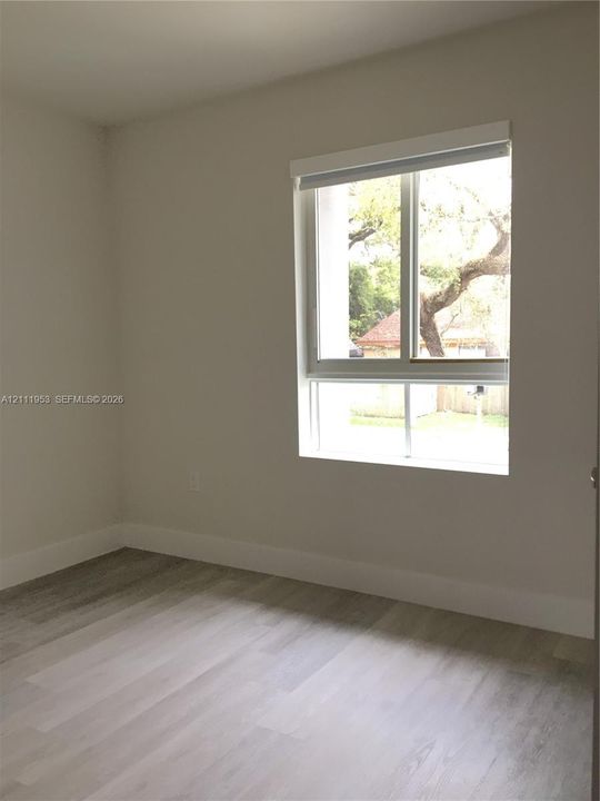 Neutral-toned interior space featuring light wood-finish flooring, crisp white wall paint, and a single-pane window with a roller shade