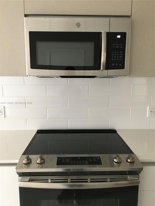 Stainless steel over-the-range microwave and electric range, white subway tile backsplash, solid-surface countertop, and flat-panel cabinetry