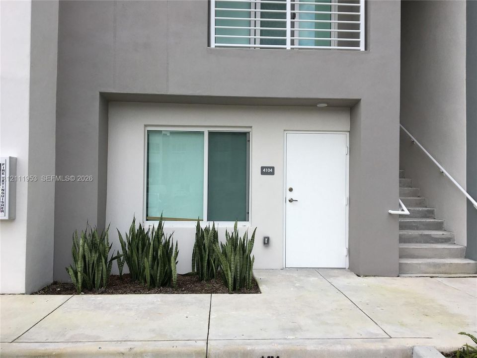 Modern exterior featuring a smooth stucco facade, a clean-lined solid entry door, concrete stairs with a metal railing, and a ground-level concrete walkway with built-in planter area