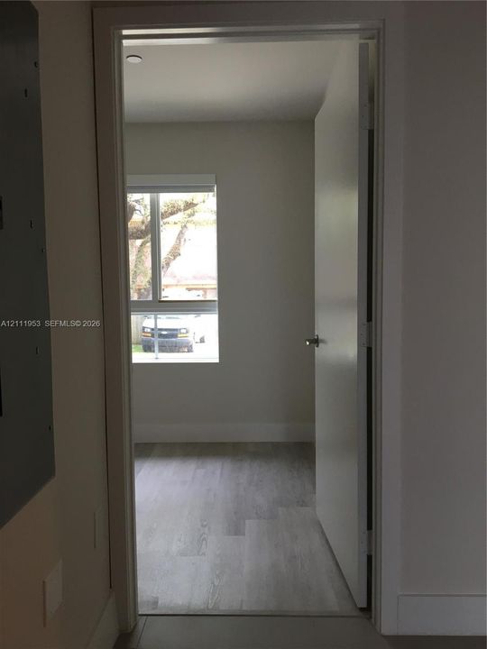Room featuring wood-finish flooring, a single-hung window with a roller shade, and a flush-panel door with a metallic lever handle