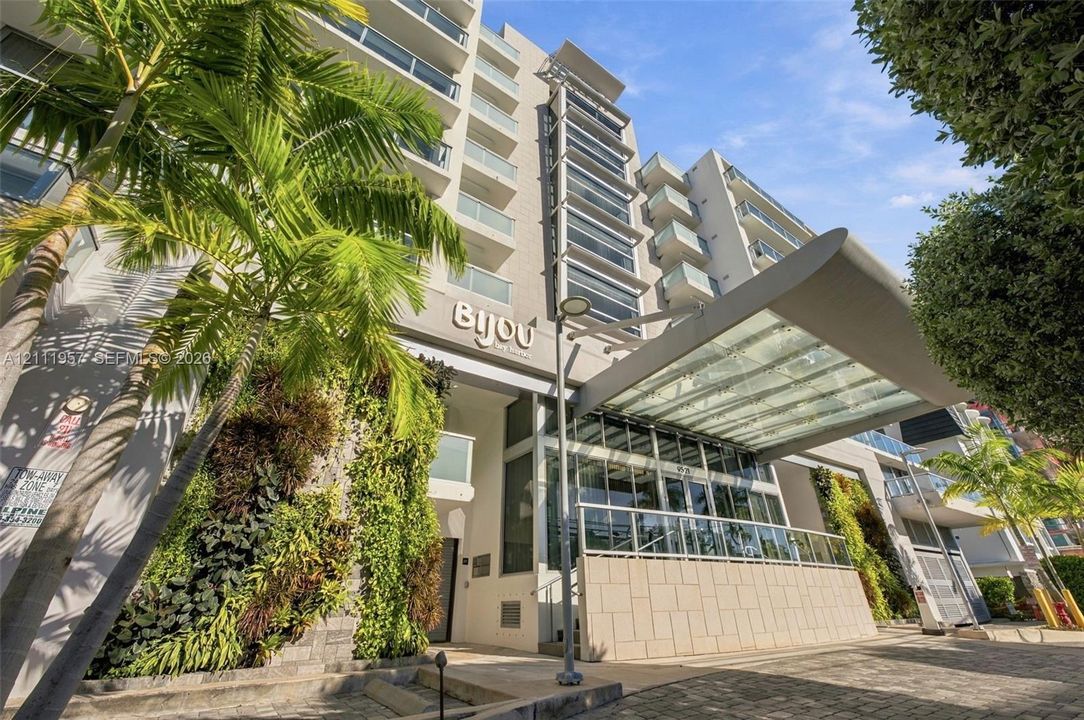 Multi-story residential building exterior featuring a glass-canopy entryway, tiered balconies with glass railings, vertical gardens, and stone-tiled facade elements