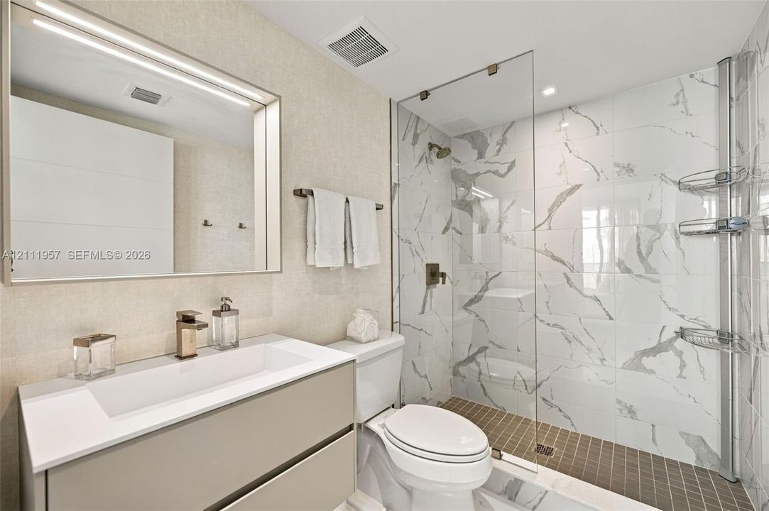 Floating vanity featuring a rectangular integrated sink, quartz countertop, and gold-tone faucet, complemented by a large framed vanity mirror, glass-enclosed walk-in shower with stone-patterned tile walls, and recessed lighting