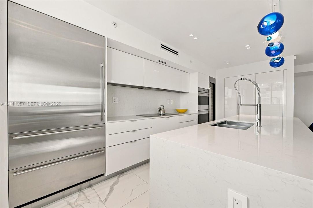 Modern kitchen featuring high-gloss cabinetry, a quartz waterfall island, stone-finish flooring, integrated stainless steel refrigeration, and a commercial-style pull-down faucet