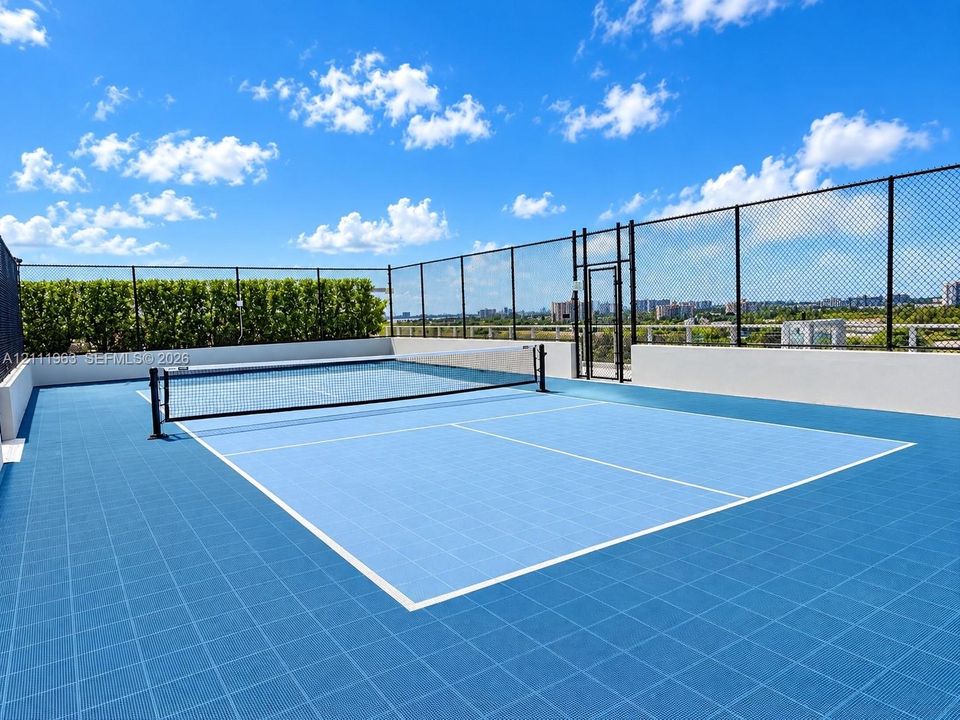 Rooftop tennis court with blue athletic tile surfacing, perimeter chain-link fencing, and integrated privacy hedges