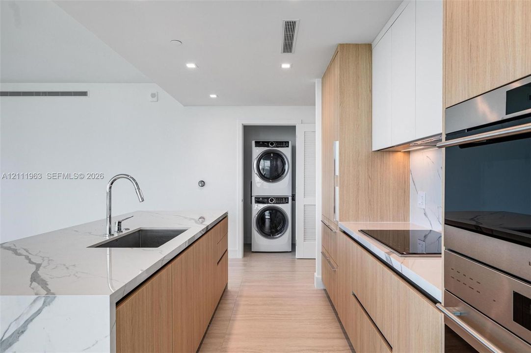 Kitchen featuring light wood-finish cabinetry, waterfall-edge stone island with an undermount sink, integrated stainless steel oven, stone backsplash, and a laundry closet with a stacked washer and dryer