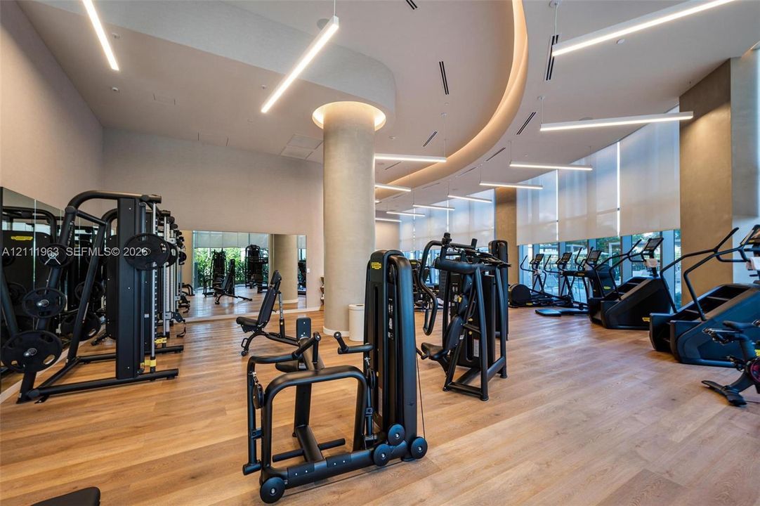 Fitness center featuring wood-finish flooring, floor-to-ceiling windows with roller shades, recessed cove lighting, suspended linear light fixtures, and a structural column