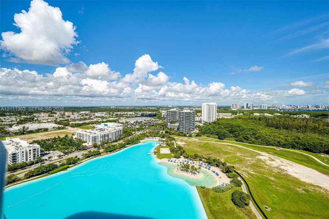 Aerial perspective featuring a large turquoise lagoon, sandy shoreline, manicured green space, and surrounding urban landscape