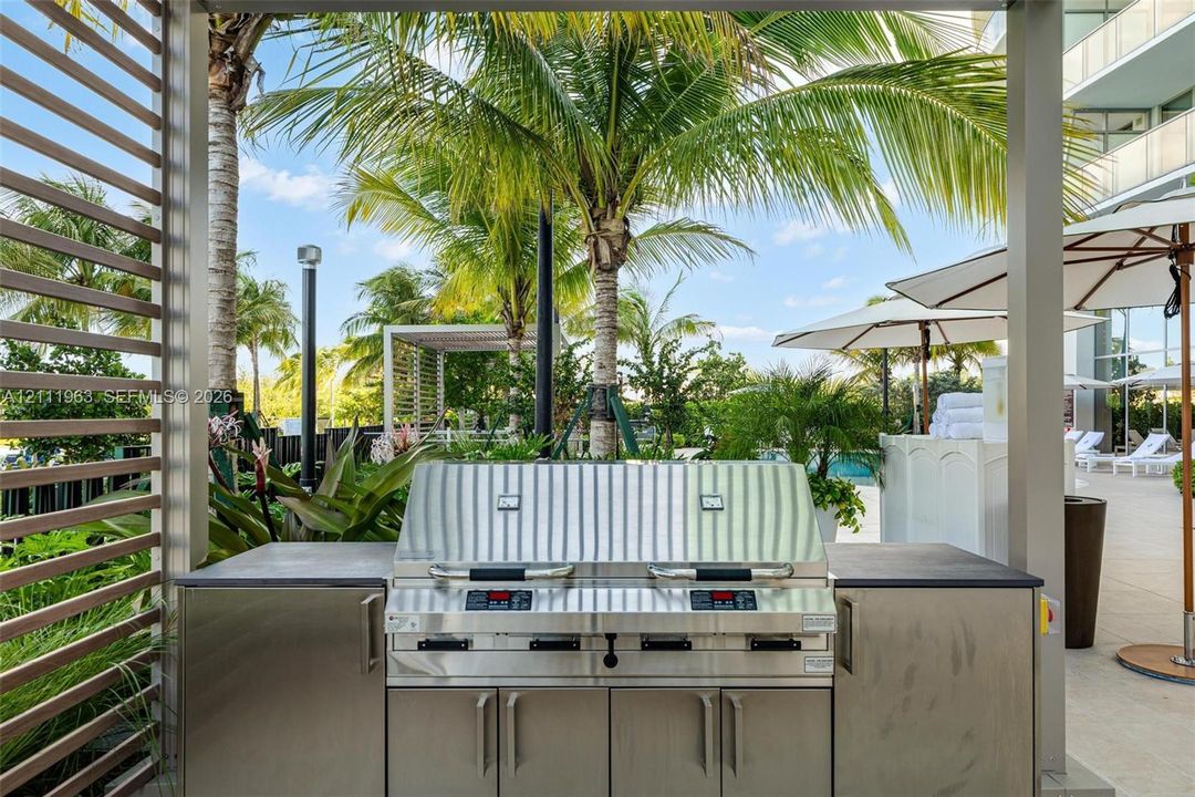 Outdoor kitchen station featuring a stainless steel built-in grill, dark stone countertop, and sleek metallic cabinetry set within a screened pavilion