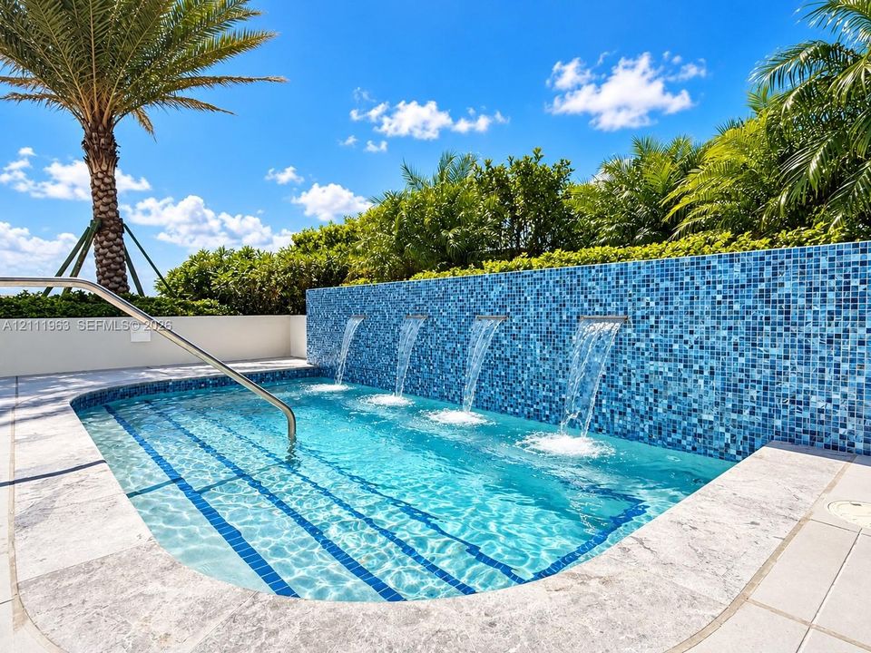 In-ground swimming pool featuring a mosaic tile water feature with four integrated spillways, stone-style pool decking, and a stainless steel entry rail