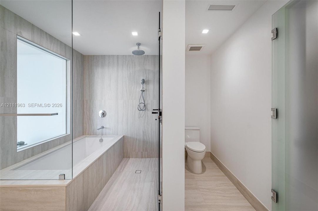 Full-height glass shower enclosure, built-in soaking tub with wall-mounted faucet, rainfall showerhead with handheld sprayer, stone-finish tile surround, and separate enclosed water closet