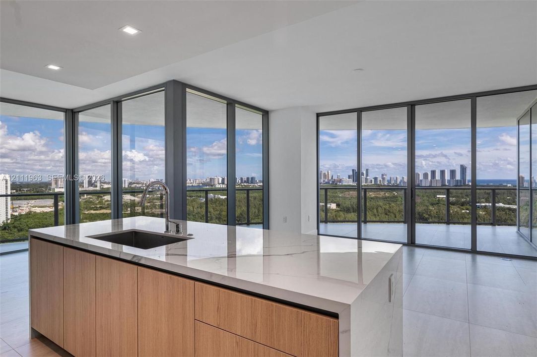 Kitchen island featuring waterfall-edge stone countertops, wood-finish cabinetry, an under-mount sink with a gooseneck faucet, floor-to-ceiling glass panel walls, and recessed lighting