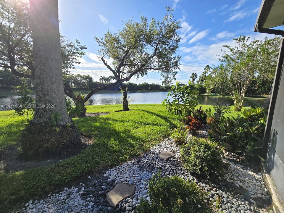 Waterfront backyard featuring a manicured lawn, mature trees, ornamental shrubs, and a decorative pebble landscape border with natural stone pavers