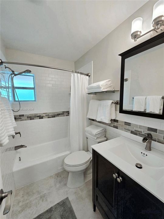 Dark-finish vanity with an integrated white sink, wall-mounted light fixture, decorative glass and stone mosaic backsplash, and shower-tub combination with white subway tile
