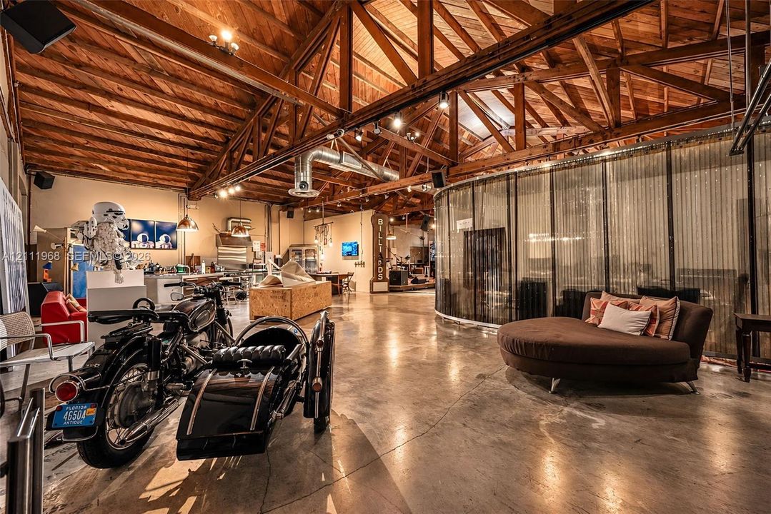Open-concept interior featuring vaulted ceilings with exposed wood trusses, polished concrete flooring, industrial metal ductwork, and a curved floor-to-ceiling metal mesh partition