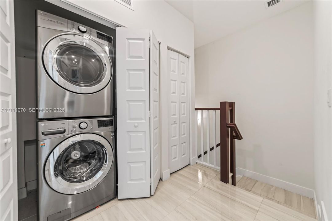 Stacked stainless steel laundry appliances, white paneled bi-fold closet doors, tile flooring, and a wooden handrail with vertical balusters
