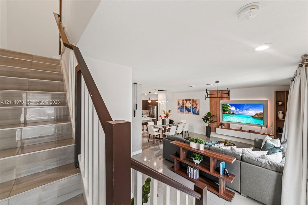 Staircase featuring wood-finish treads, a dark-toned handrail, and white vertical balusters, opening into a living area with large-format tile flooring, recessed lighting, and a custom built-in entertainment center with accent lighting
