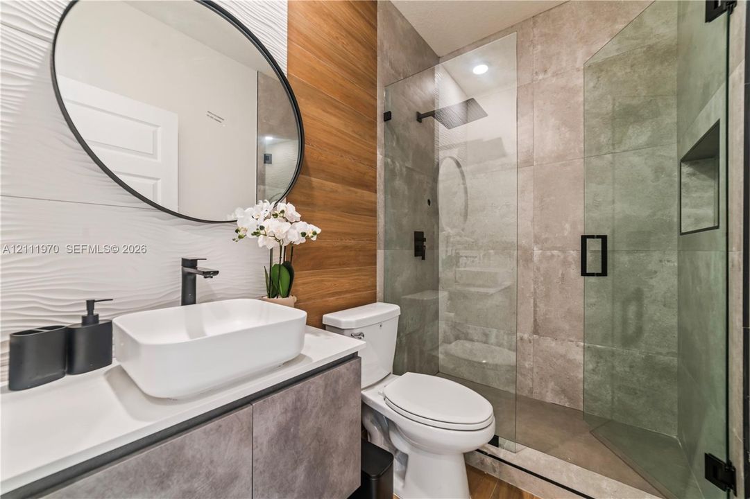 Bathroom featuring a wall-mounted vessel sink, matte black fixtures, textured wave-patterned tile wall, wood-finish paneling, and a glass-enclosed walk-in shower with large-format tile surround