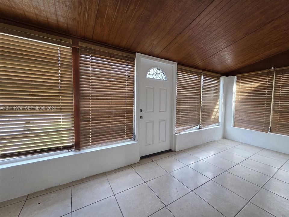 Enclosed porch featuring wood-paneled ceiling, large-format tile flooring, multiple windows with fitted blinds, and a white entry door with a decorative arched transom window