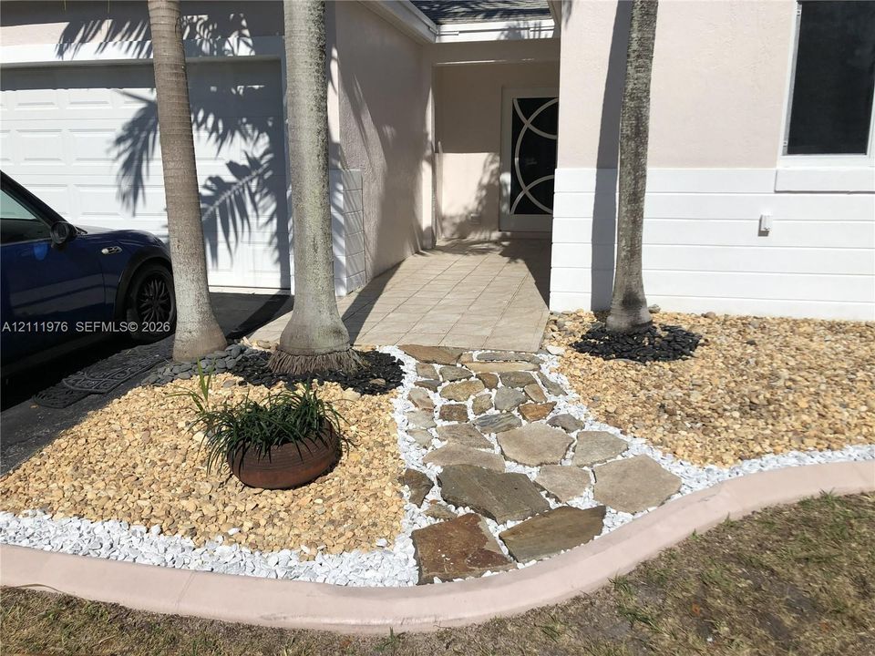 Exterior entrance featuring a flagstone walkway, multi-textured stone landscaping, palm trees, and a decorative circular-pattern front entry door