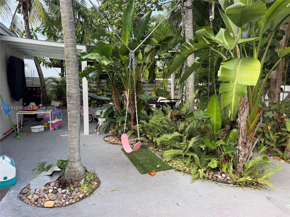 Concrete patio featuring a covered area, mature palm trees, tropical landscaping, and river rock garden beds