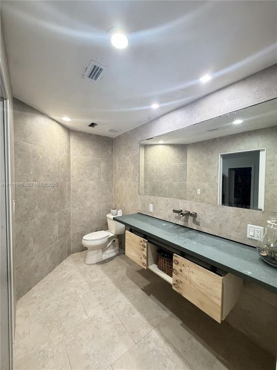 Floating wood-finish vanity with a dark solid-surface countertop and wall-mounted faucet, complemented by full-height tile wall cladding and recessed ceiling lighting