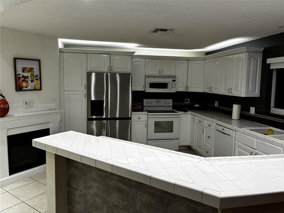 Kitchen featuring white cabinetry, a peninsula with tile countertops, built-in stainless steel appliances, and a fireplace with a white mantel