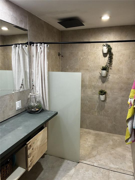 Bathroom featuring a stone-finish tile surround and flooring, a matte black rainfall shower head, a frosted glass shower partition, and a vanity topped with a dark surface