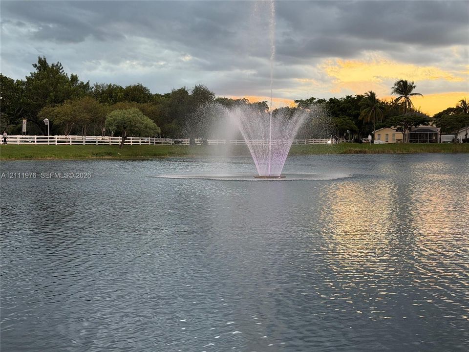 Large lake featuring a central illuminated water fountain, manicured grassy embankment, and white perimeter fencing