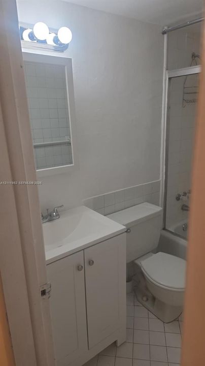 Bathroom featuring a white vanity cabinet, ceramic tile flooring, a shower-tub combination with a glass enclosure, and a vanity-mounted mirror with integrated light fixture