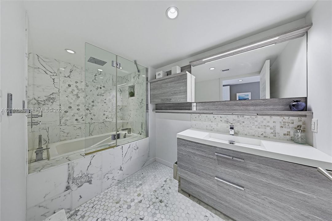Bathroom featuring a wood-finish floating vanity with an integrated sink, a glass-enclosed soaking tub with stone-tiling, a wall-to-wall mirror, and hexagonal tile flooring