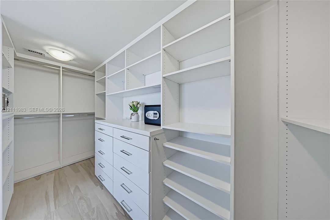 Walk-in closet featuring extensive built-in white cabinetry, multiple tiered shelving units, integrated drawer storage, and light-tone wood-finish flooring