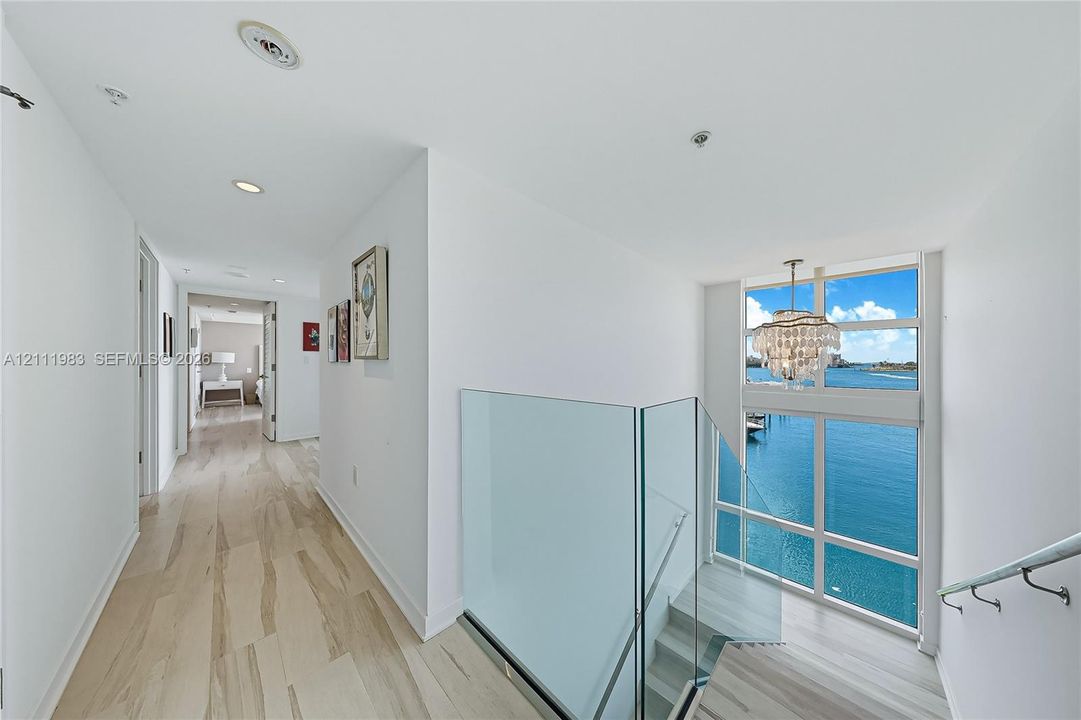 Upstairs hallway and staircase featuring light-toned wood-finish flooring, glass stair railings, multi-panel floor-to-ceiling windows, and a decorative chandelier